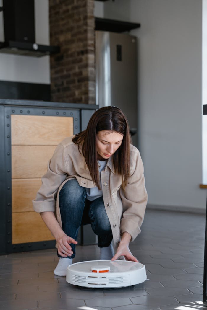 who-we-are Woman adjusting a robotic vacuum cleaner on a tiled floor indoors.